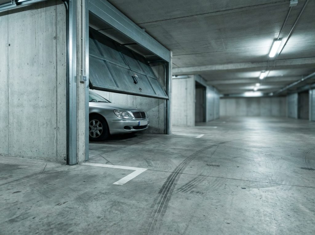 Box de parking souterrain en béton avec porte métallique entrouverte et voiture garée à l'intérieur